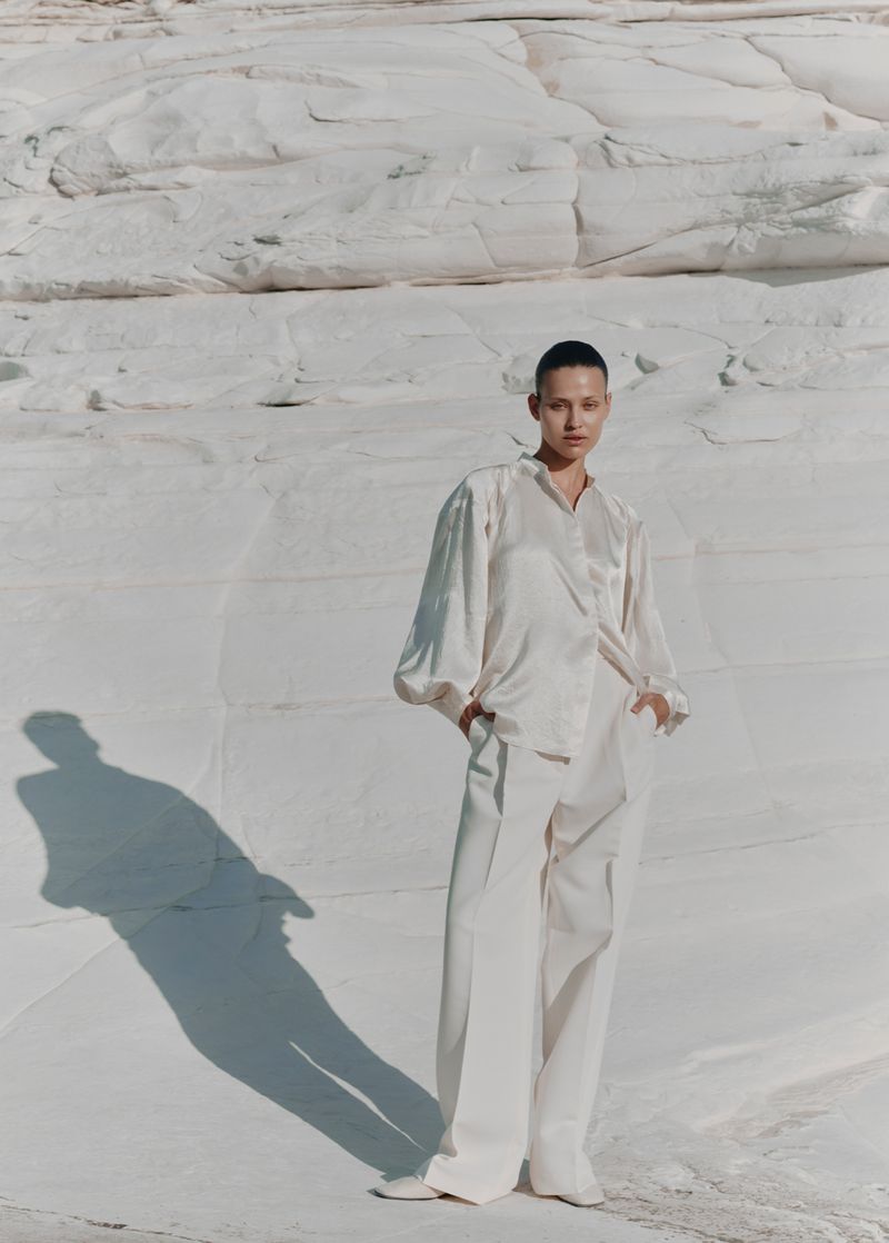 Woman in a monochromatic off-white outfit standing on a white rock formation, posing with a relaxed posture, and her shadow visible.
