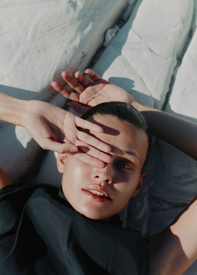 Portrait of a model lying down with one hand covering the eye, wearing black top against white bedsheets in soft, natural light.
