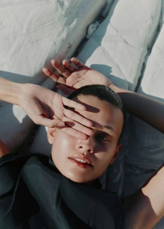 Portrait of a model lying down with one hand covering the eye, wearing black top against white bedsheets in soft, natural light.