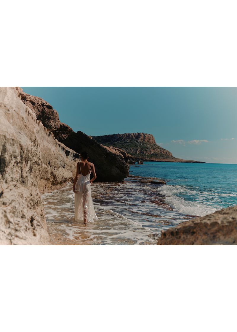 Woman in white dress walking on rocky beach towards the ocean with clear blue sky and turquoise water on a sunny day.