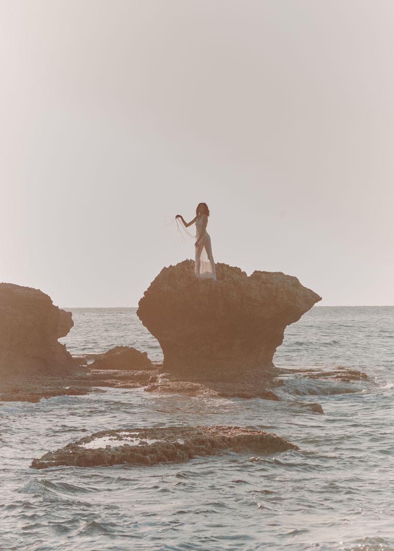Woman in sheer dress stands on a rock formation in the ocean at sunset, ocean waves, and horizon.