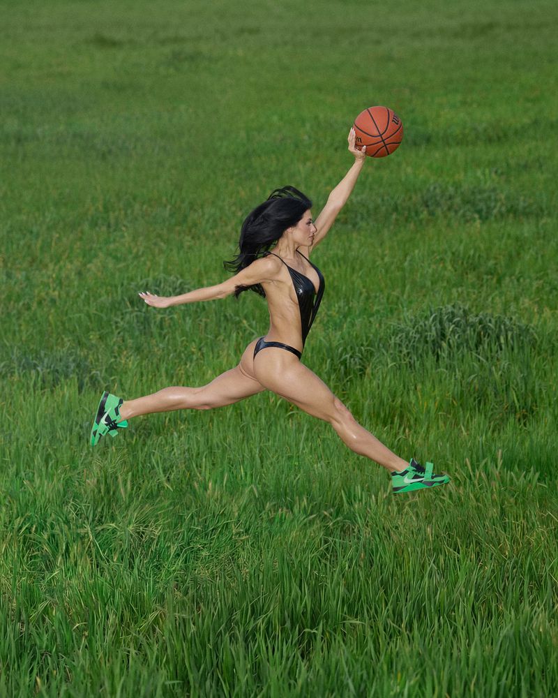 Woman in a black bodysuit leaping across a green meadow while holding up a basketball and wearing green sneakers.