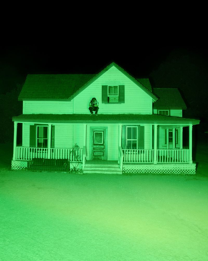 Person seated on the roof of a two-story white house at night under a distinct green monochromatic night-vision camera lighting effect.