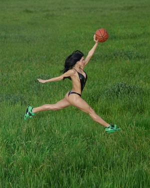 Woman in a black bodysuit leaping across a green meadow while holding up a basketball and wearing green sneakers.