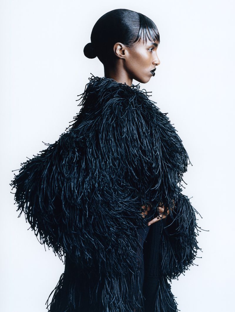 Profile of a woman with dark hair and lipstick wearing a coat with long, thin, fringe-like strands, set against a bright white background.
