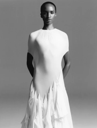 Elegant black woman in flowing white gown poses in a studio against a grey backdrop in a portrait-style shot. Fashion editorial.
