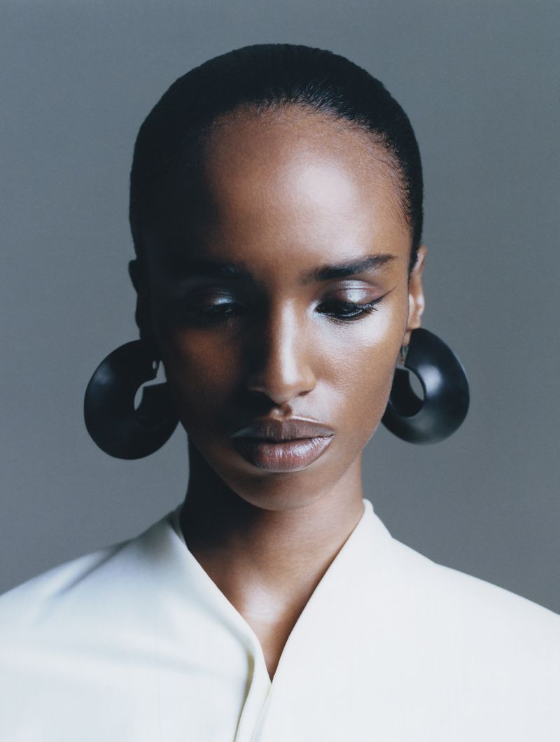 Close-up studio portrait of a Black woman with smooth skin, slicked-back hair, large black disc earrings, and a light-colored top.
