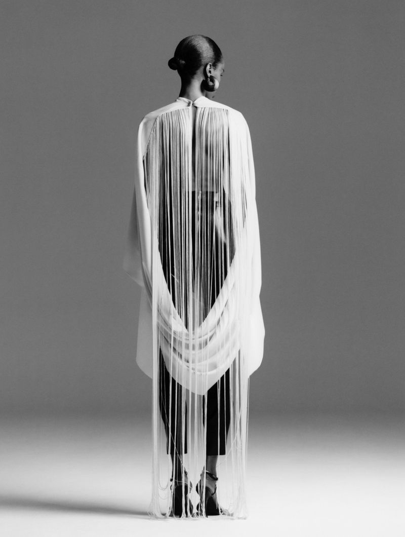 Bibi Abdulkadir viewed from behind wearing a white garment with flowing floor-length fringe against a grey studio backdrop.