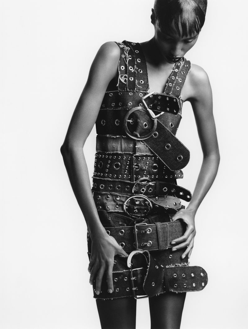 Monochrome fashion shot featuring a model wearing a dress made of belts and grommets against a bright, neutral background in a studio.