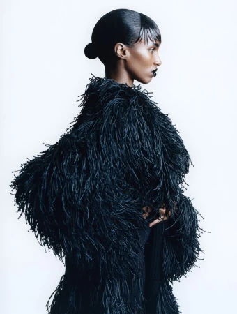Model Bibi Abdulkadir in profile wearing a voluminous black fringe coat and sleek hairstyle against a white backdrop.