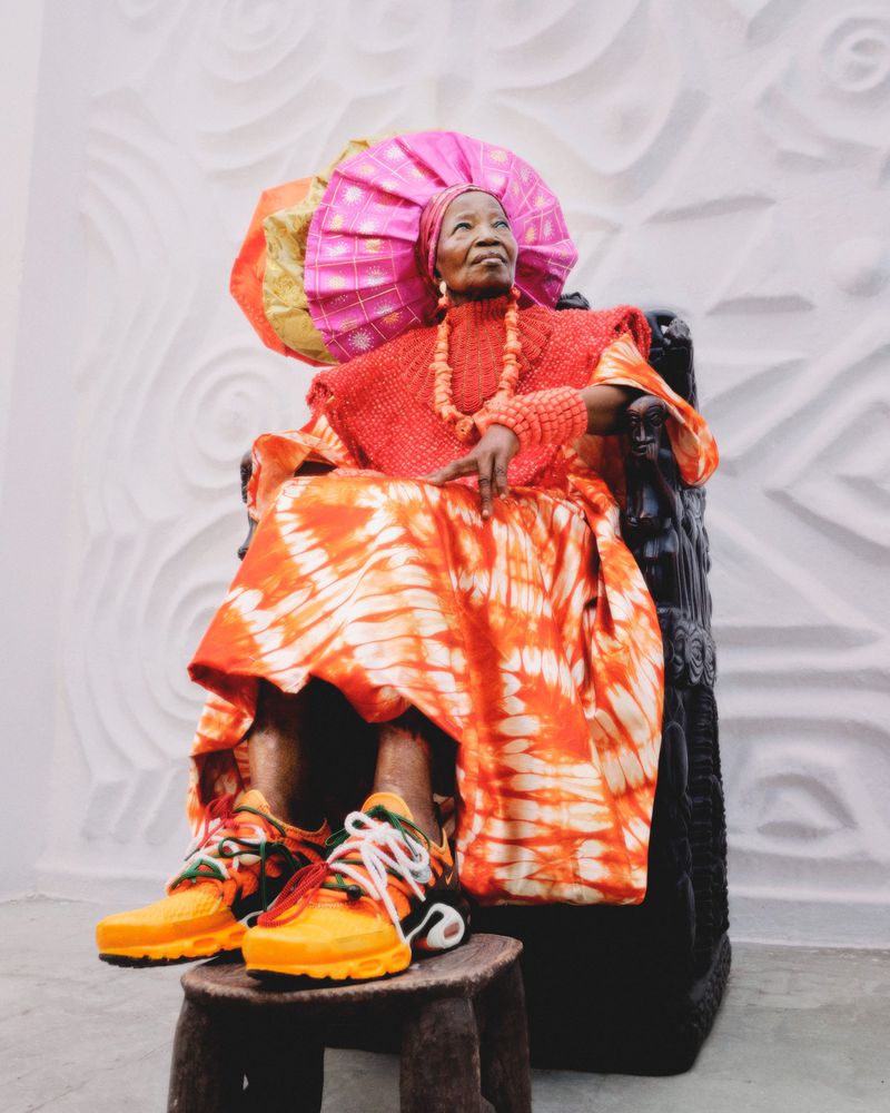Elderly woman wearing a vibrant patterned orange dress and large pink headwrap, seated on a carved chair with yellow sneakers.