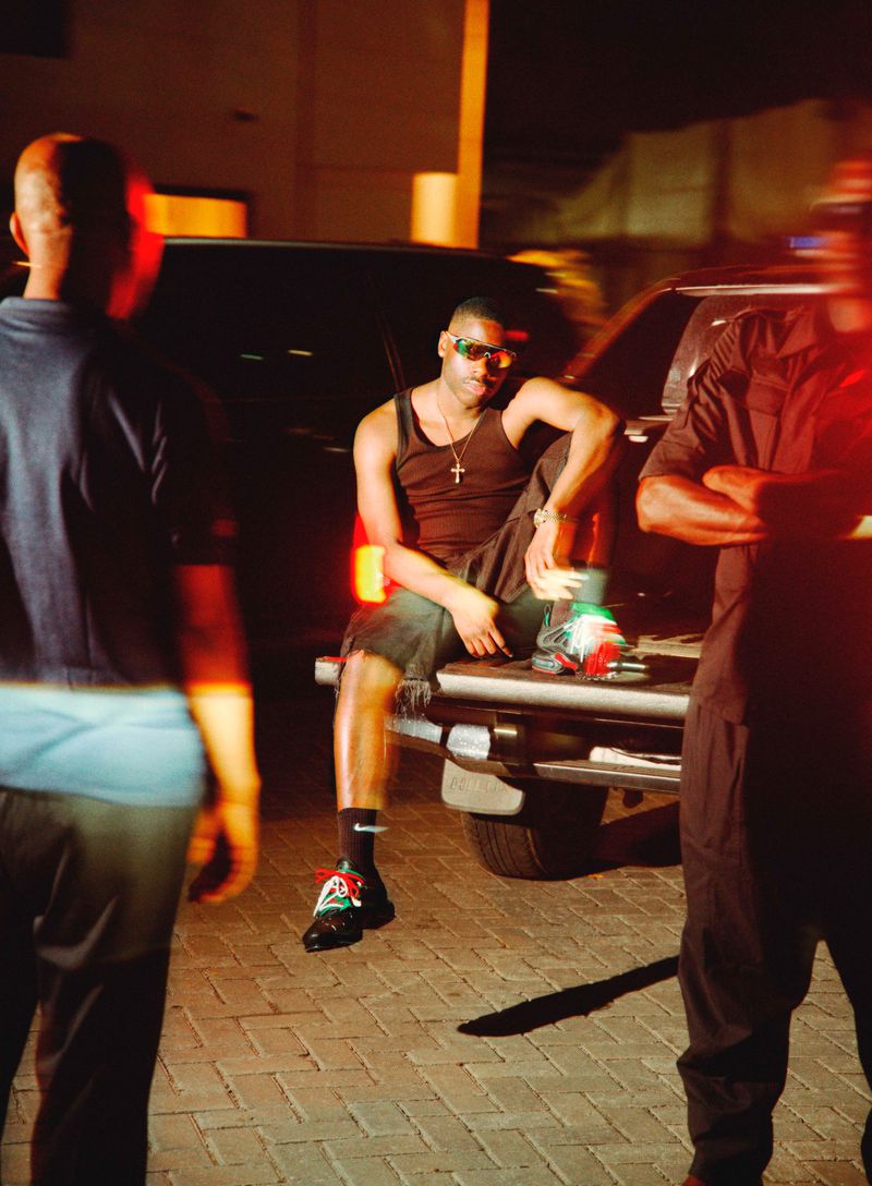 Black man in a black tank top and shorts seated on the back of a pickup truck at night with out-of-focus figures in the foreground.