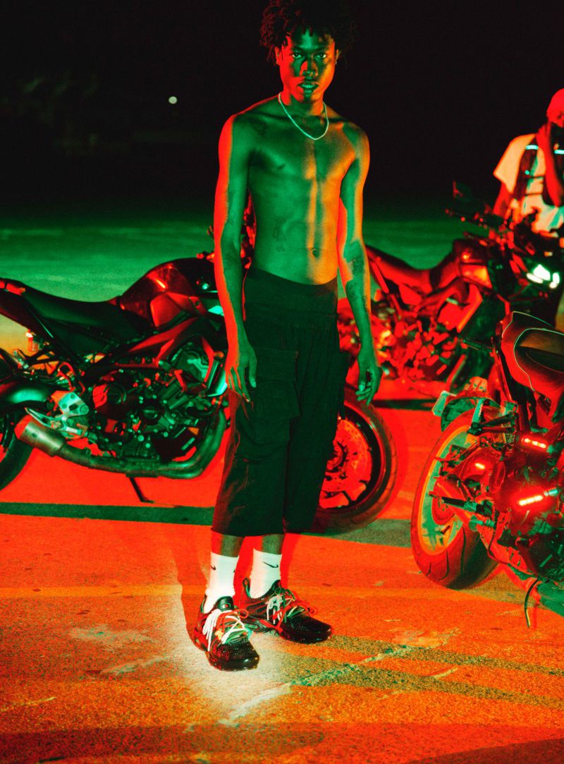 Shirtless man standing outdoors at night between two motorcycles under dramatic red and green lighting