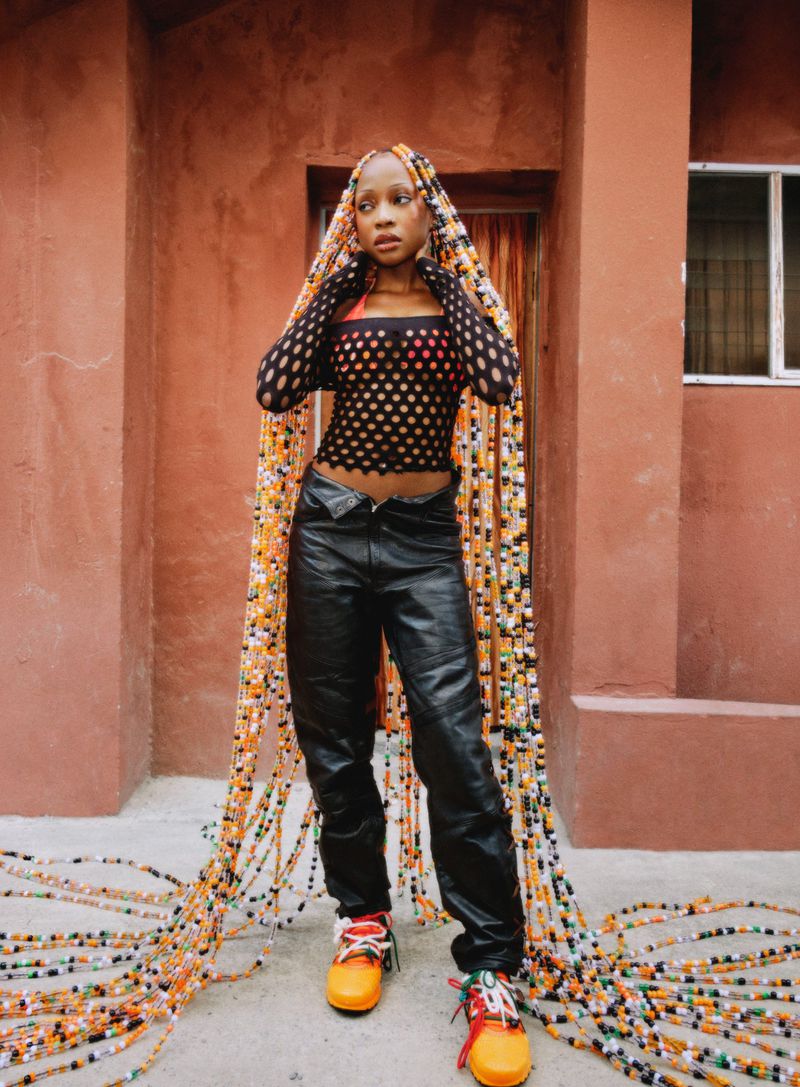 A woman wearing black leather trousers, patterned mesh top, and long braided hair pooling onto the ground outside a terracotta building.