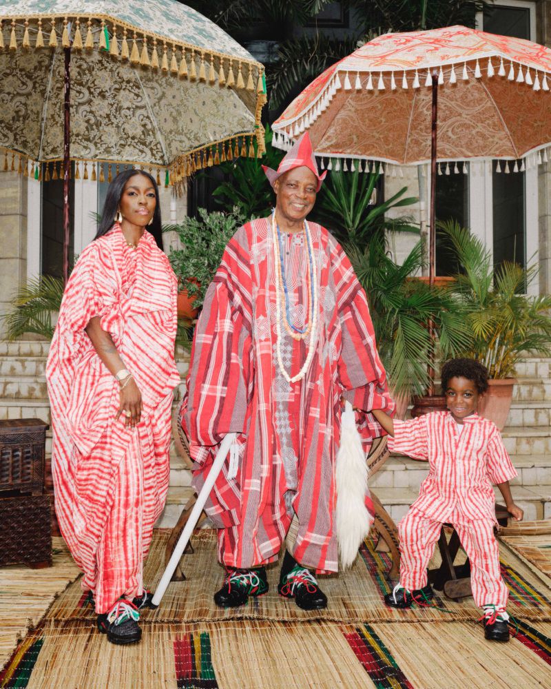 A family group stands on a mat in front of a building, wearing red and white striped outfits and traditional hats, with umbrellas overhead.