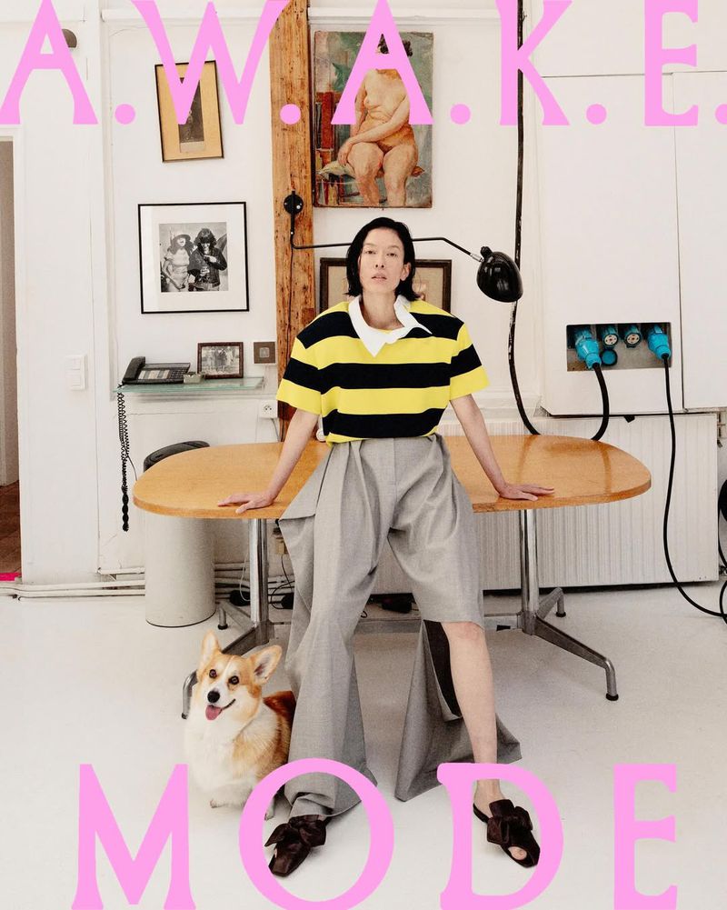 Model wearing yellow and black striped polo and grey trousers sitting on a wooden table with a corgi below, framed by pink lettering