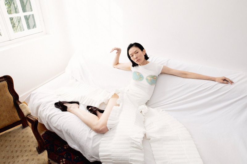 Model lying on a white covered sofa wearing a white top and pleated skirt for A.W.A.K.E. MODE S/S 2026 campaign