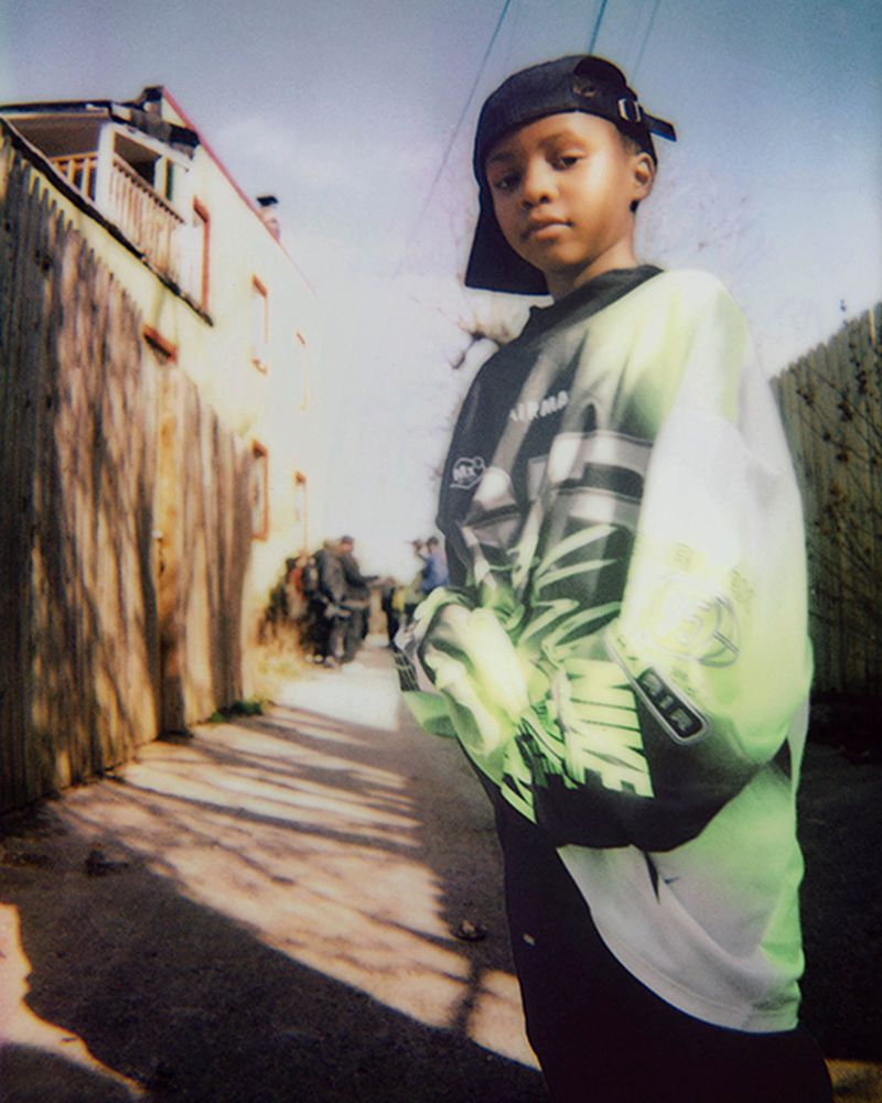 Young person in cap and 90s-style patterned shirt stands on sunlit street with buildings, wall, and other people in background.