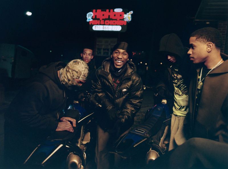 Group of young Black men gathered around motorcycles at night outside a lighted storefront in Baltimore.