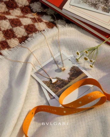 Elevated view of gold Bulgari necklaces on top of photographs, white fabric, ribbon, daisies, and books; still life jewelry product shot.