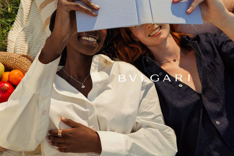 Two smiling women holding an open book over their faces outdoors, wearing necklaces and rings for Bulgari