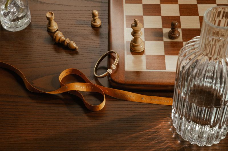 A Bulgari gold bracelet and branded ribbon resting on a dark wood table with a chessboard and glass vase.