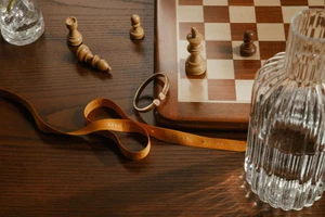 A Bulgari gold bracelet and branded ribbon resting on a dark wood table with a chessboard and glass vase.