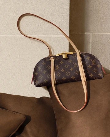 Monogram canvas Louis Vuitton shoulder bag with leather straps resting on a brown cushion against a stone wall