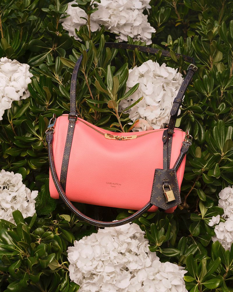 Coral pink Louis Vuitton shoulder bag with monogram straps resting among lush green leaves and white flowers