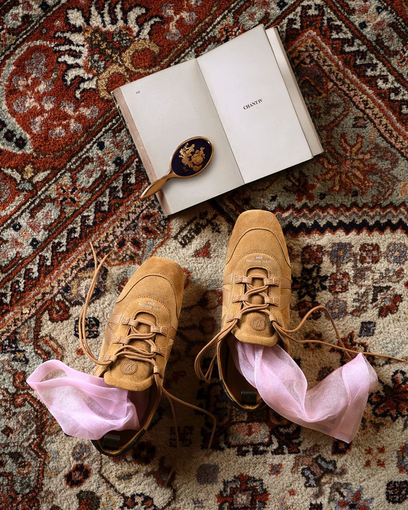 Tan suede sneakers with light pink sheer socks tucked inside, placed on an ornate patterned rug beside an open book and small vintage hand mirror.