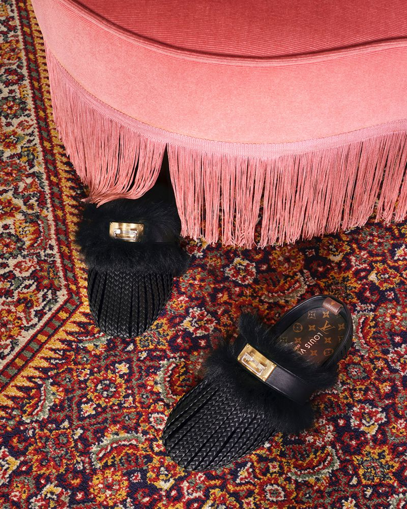 Black furry slip-on shoes with gold hardware beneath a pink fringed stool on a patterned carpet
