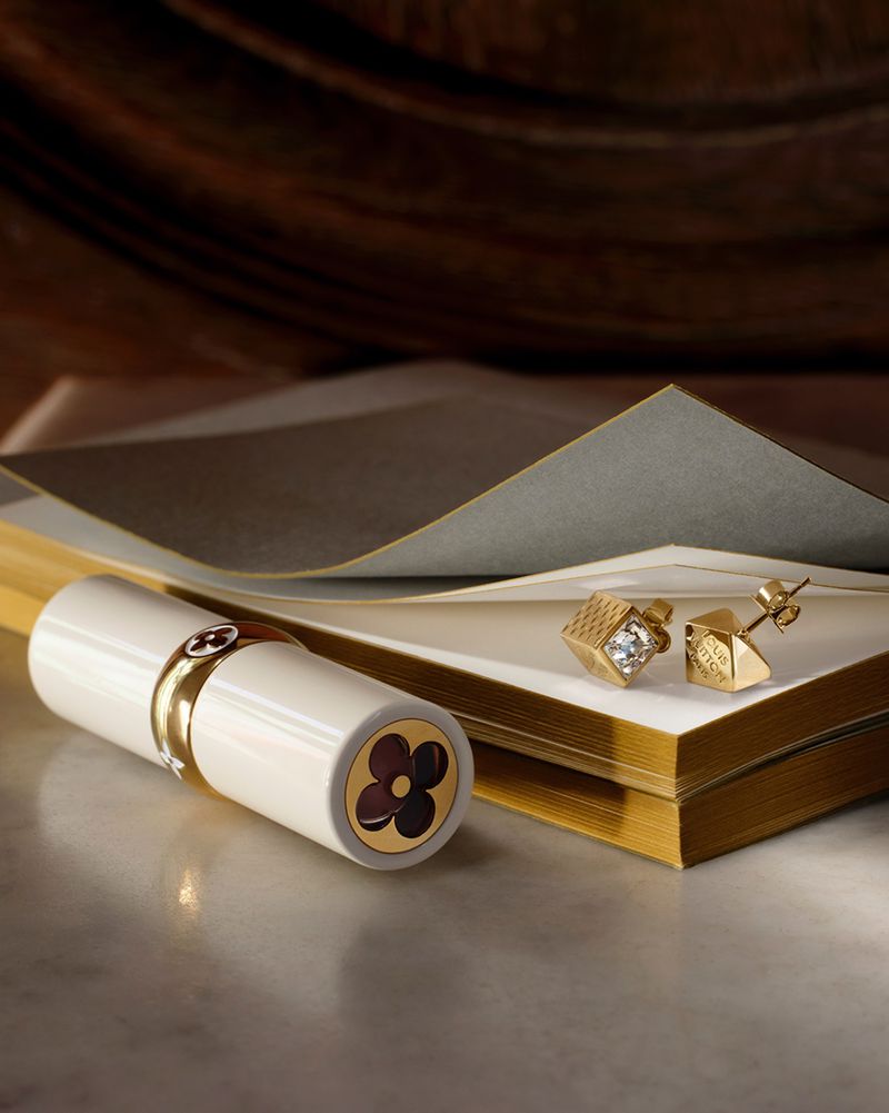 Close-up product shot of a white lipstick tube and gold earrings resting on a stack of cards with gold edges