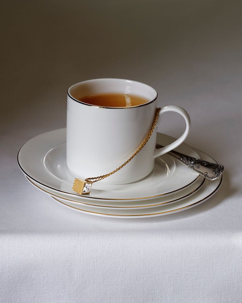 White teacup on stacked saucers with a gold chain necklace draped over the rim resting on a white tablecloth