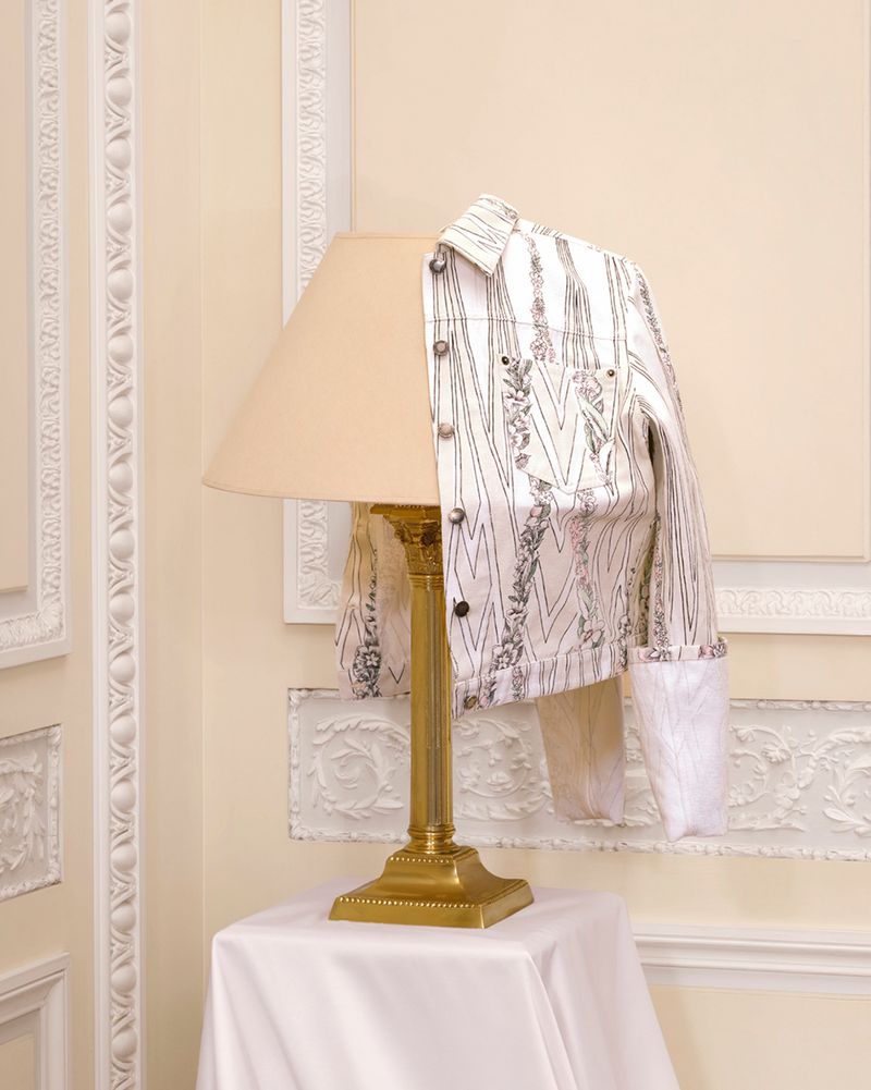 A patterned white jacket draped over a table lamp with a beige shade against a pale wall with decorative white molding.