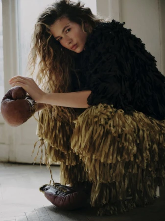 Model with long curly hair wearing a textured black top and fringed skirt while holding brown leather boxing gloves indoors.