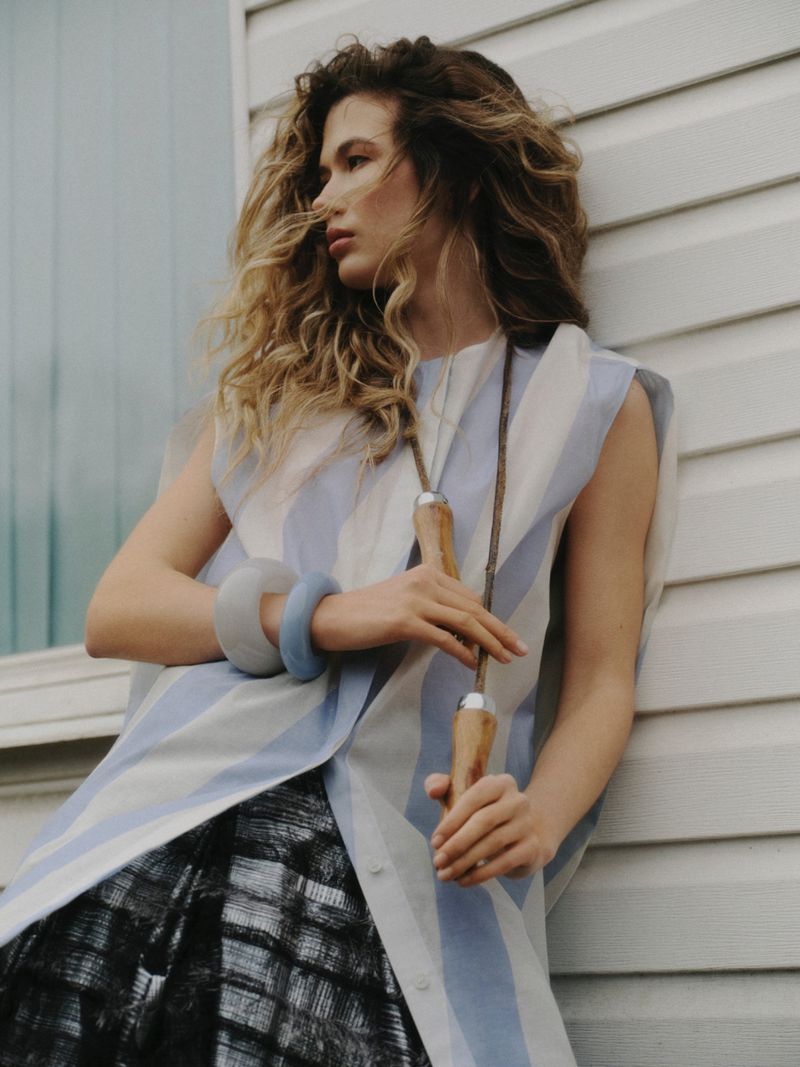 Model Meg Dmitruk wearing striped top and holding wooden tool leaning against white exterior wall for Elle Poland