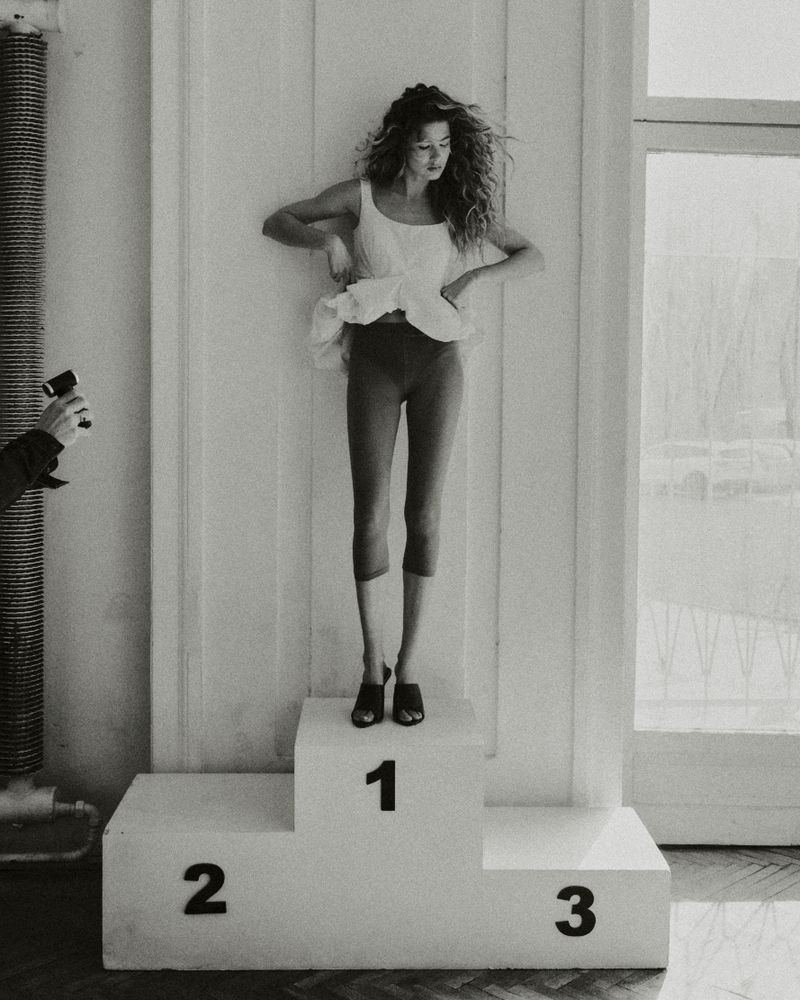 Model standing on a first-place podium wearing a white tank top and dark leggings for an Elle Poland editorial shot by Dorota Szulc.