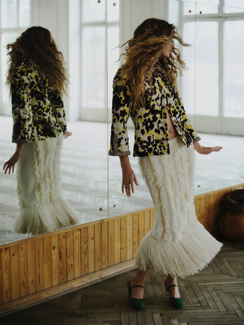 Model with long wavy hair wearing a patterned jacket and fringed white skirt posing in front of large mirrors.