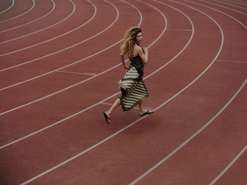 A female model running on a red running track wearing a striped dress and sandals, captured from a high-angle perspective.