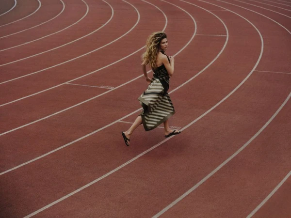 A female model running on a red running track wearing a striped dress and sandals, captured from a high-angle perspective.