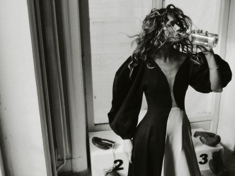 Model drinking from glass bottle wearing a black and white gown with voluminous sleeves for Elle Poland editorial