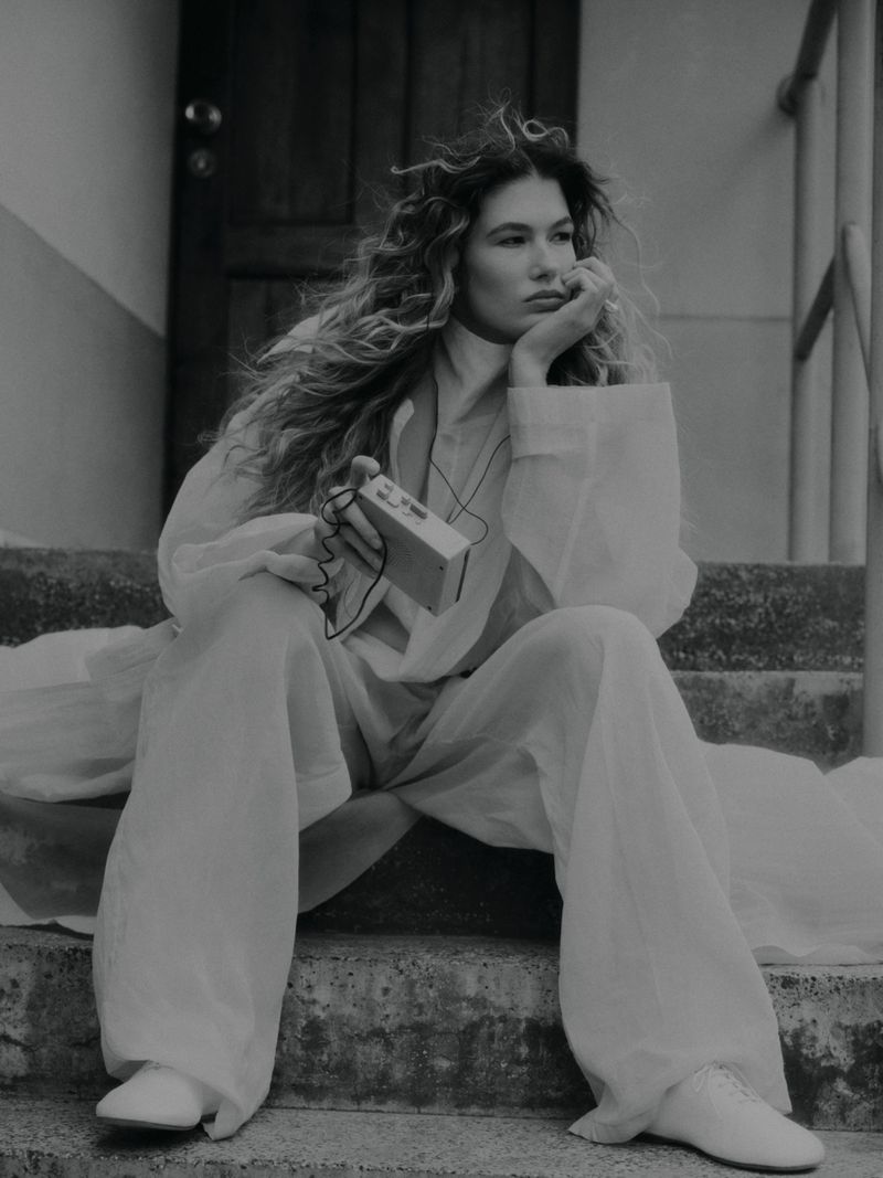 Model with long curly hair seated on stone steps holding a small portable audio player, wearing a light-coloured loose shirt and trousers.