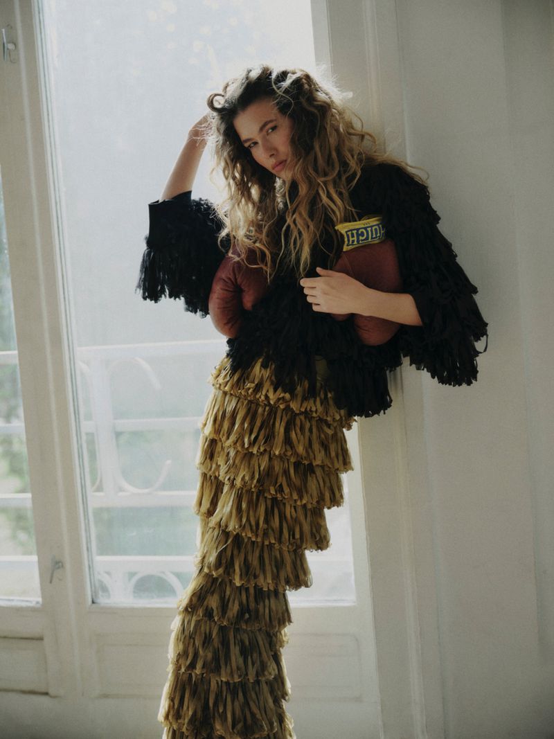 Model Meg Dmitruk wearing a fringed black top and tiered gold skirt with boxing gloves by a window