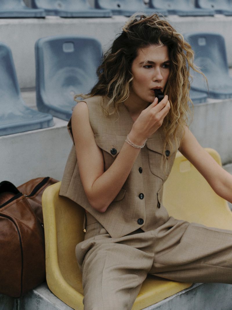 Model seated on yellow stadium seats wearing a tan vest and trousers while holding a small black whistle.