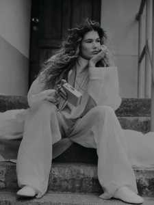 Model with long curly hair seated on stone steps holding a small portable audio player, wearing a light-coloured loose shirt and trousers.