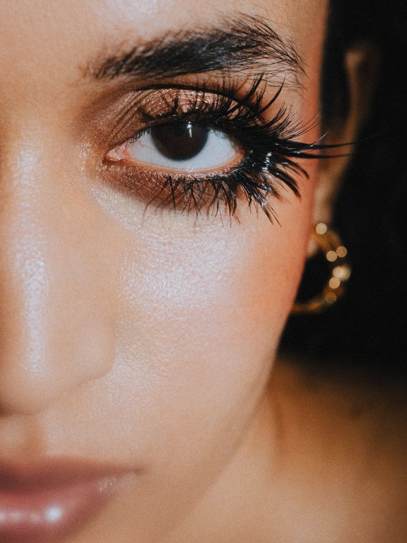Extreme close-up beauty portrait showing dramatic spidery black eyelashes, shimmery bronze eyeshadow, and a gold hoop earring.