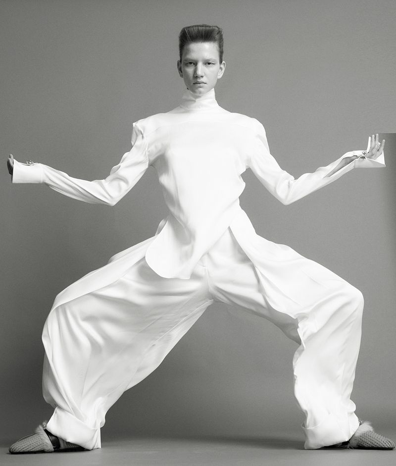 Model in a white high-neck top and wide-leg trousers posing dynamically against a grey seamless background.