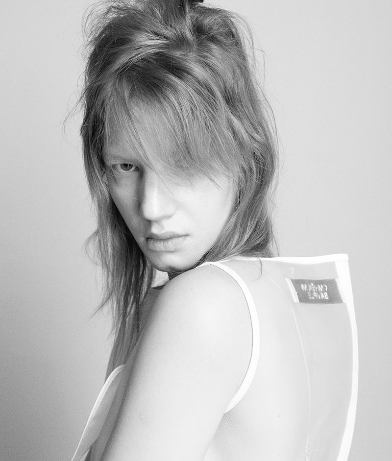 Close-up portrait of a female model with messy blonde hair wearing a white top against a neutral background.