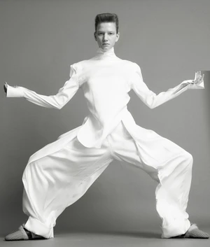 Model in a white high-neck top and wide-leg trousers posing dynamically against a grey seamless background.