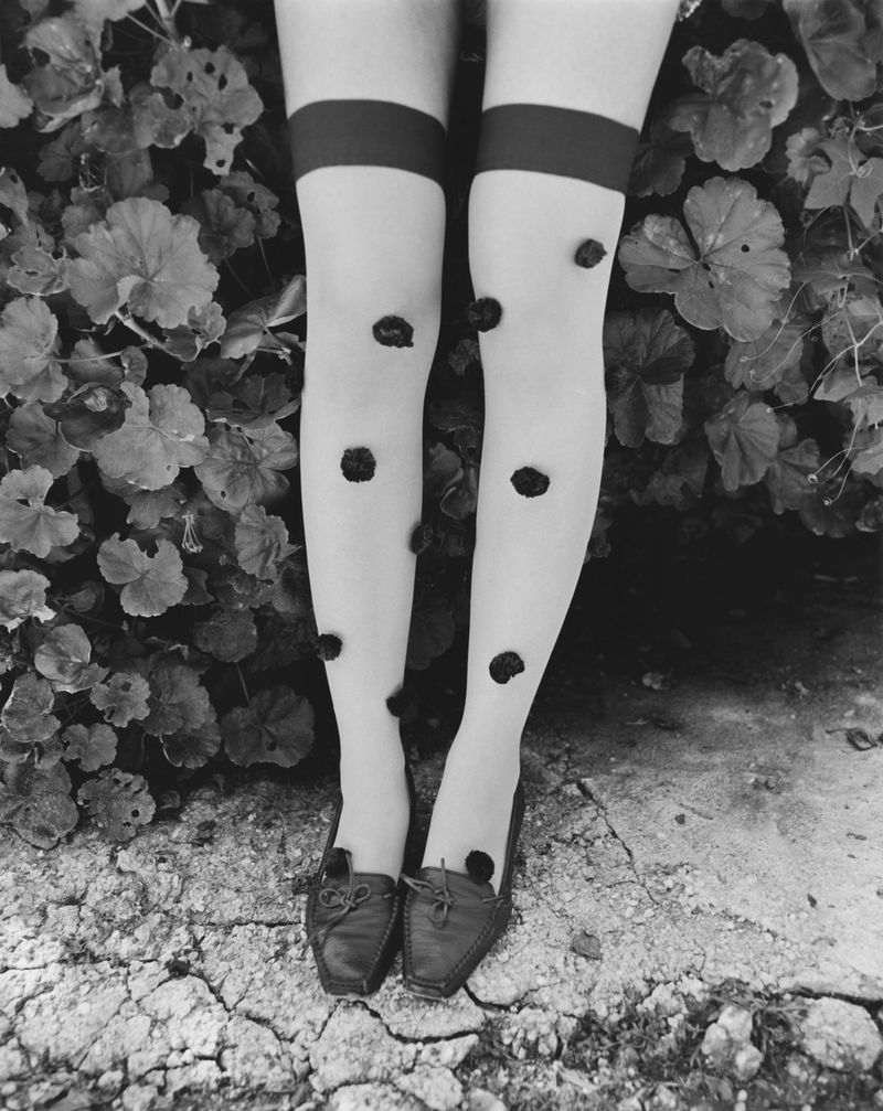 Monochrome image of a person's legs wearing polka-dot tights, dark-colored loafers, standing in front of foliage and a cracked surface.